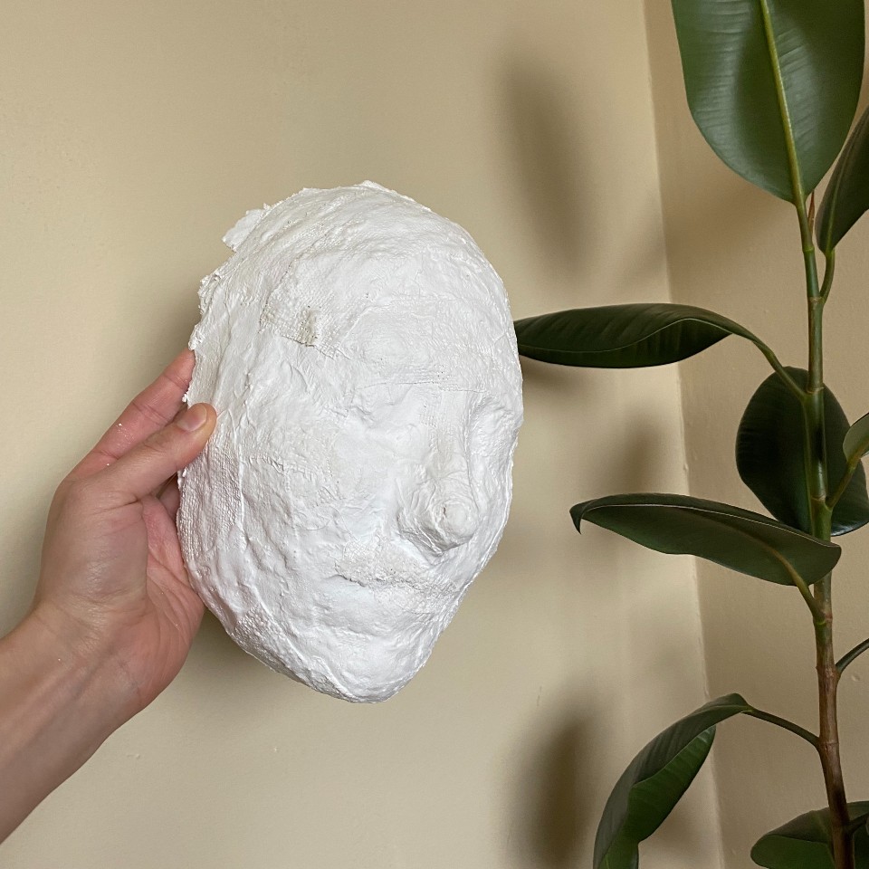 Photo of Shion Skye Carter holding aloft a white plaster mask of her face with Ficus plant in the corner